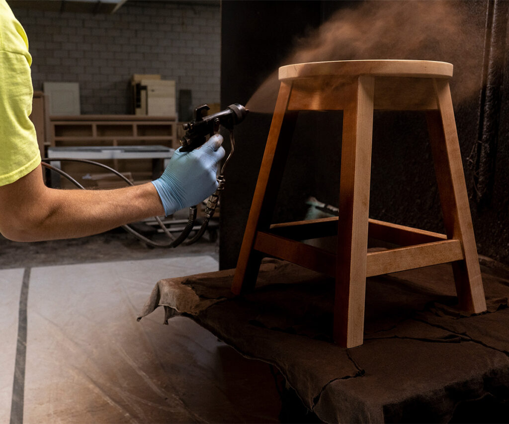 Worker spraying stain and topcoat on hardwood stool for color and long-term protection.
