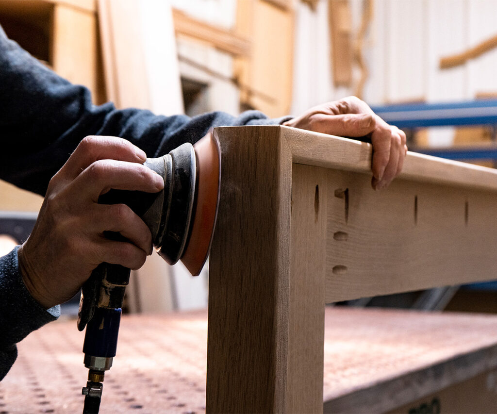 Craftsman sanding hardwood furniture corner to refine edges and smooth the surface.